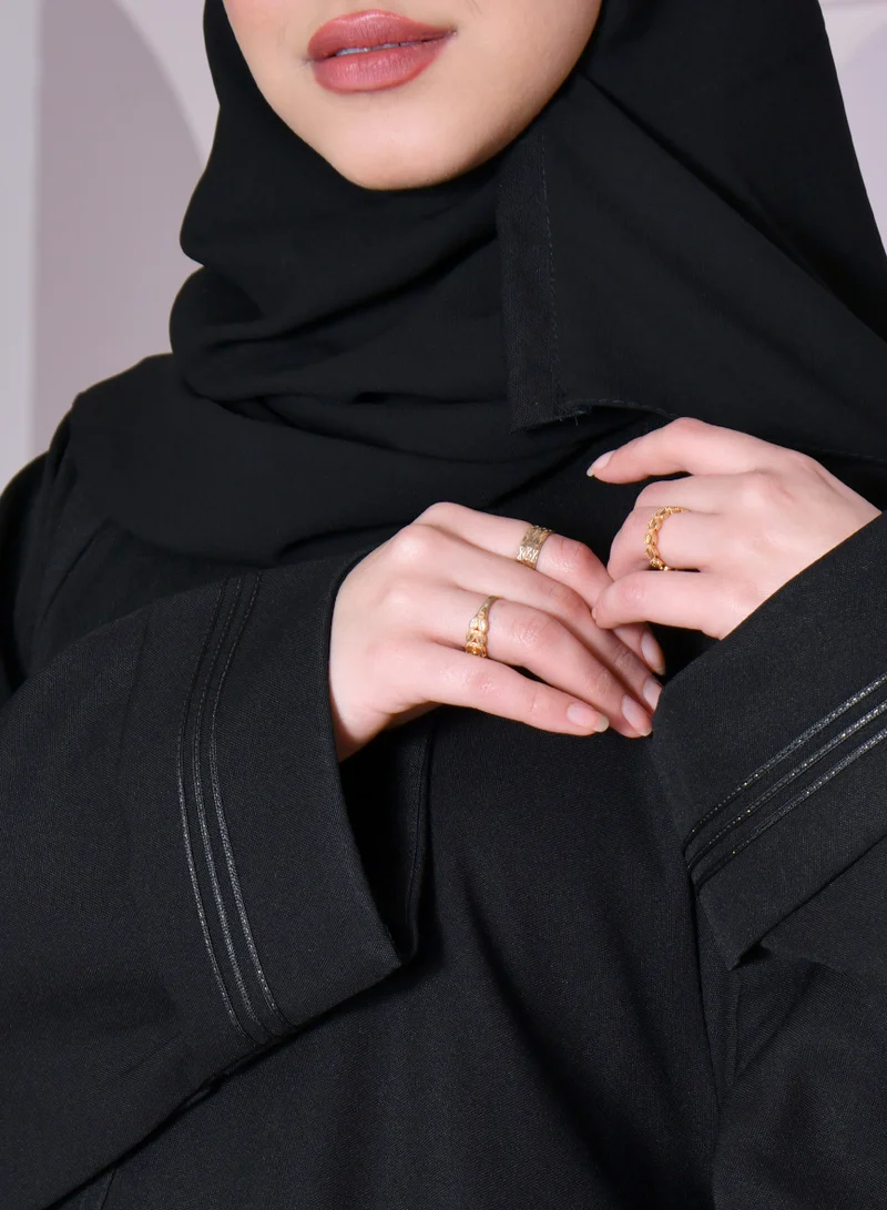 HAWRAA ABAYA A black, closed abaya with a wide, bell-shaped cut and delicate gold embroidery on the sleeves and lower hem.