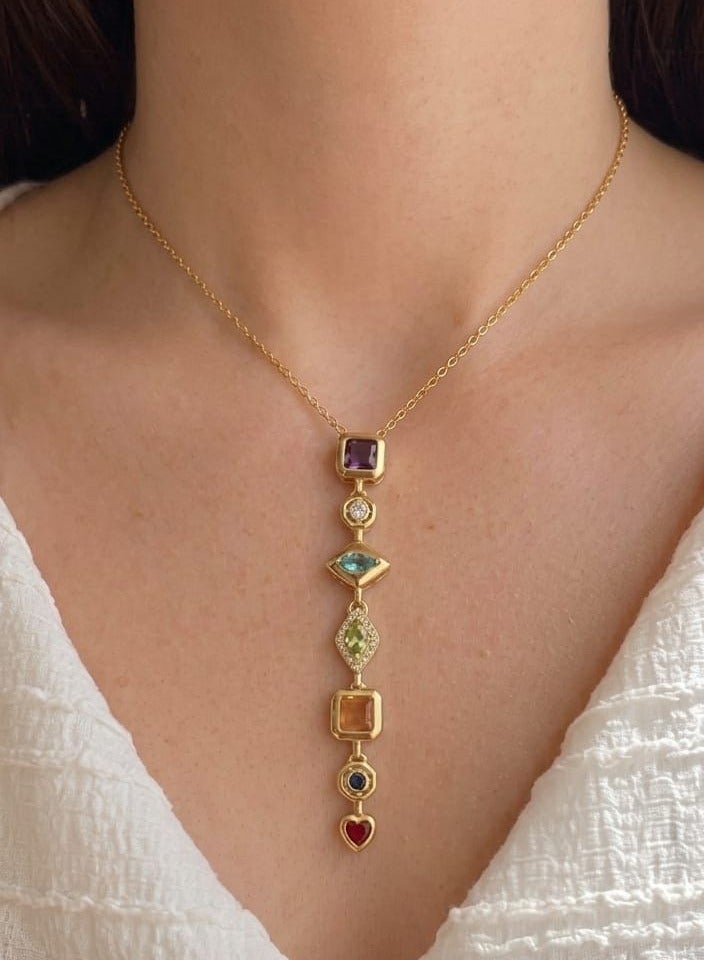 Luxuria An elegant gold-plated pendant necklace, featuring a longitudinal design studded with seven gemstones of various shapes and colors, arranged vertically to end with a small red heart-shaped pendant. - Image 1