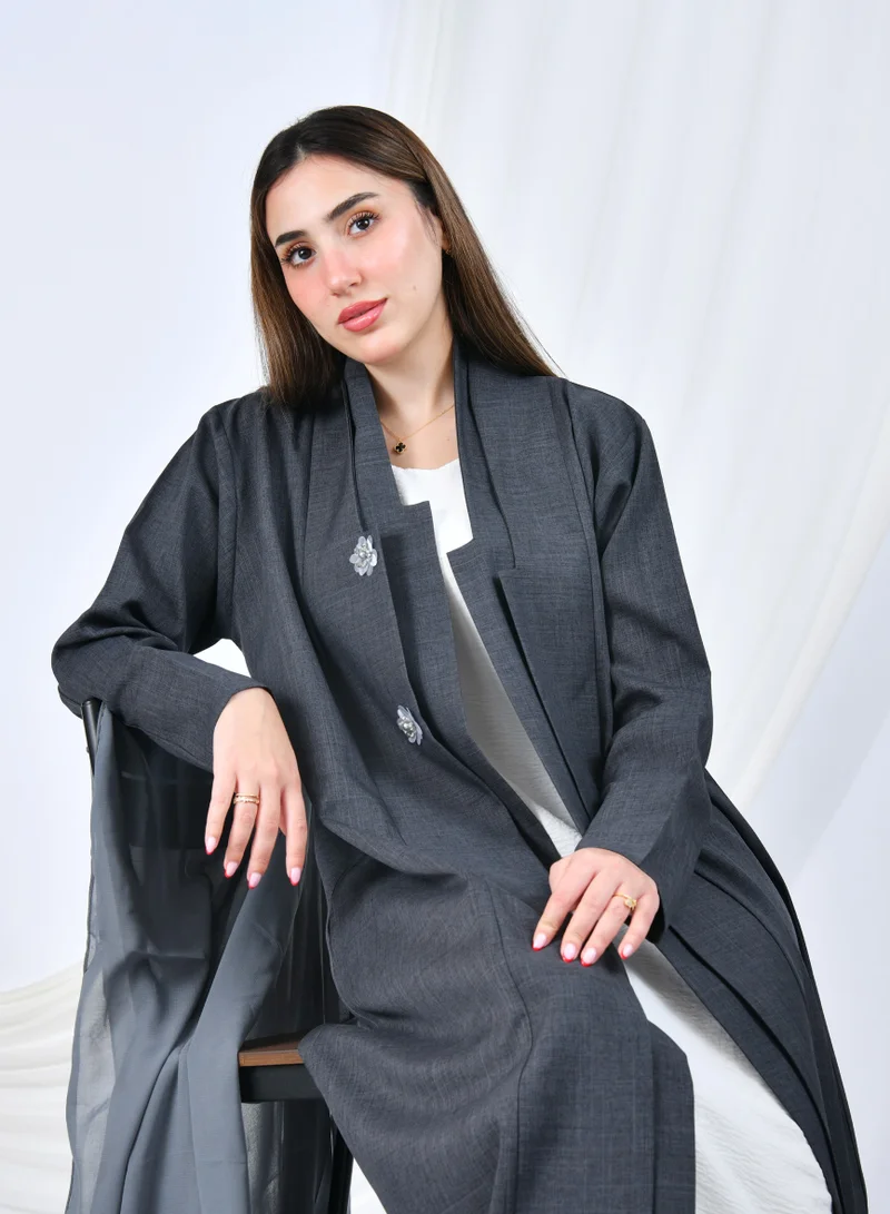عبايات حوراء عباية رسمية باللون الرمادي بتصميم بسيط وقصة ربع كلوش، مزينة بأزرار على شكل زهرة تضيف لمسة أنثوية راقية.