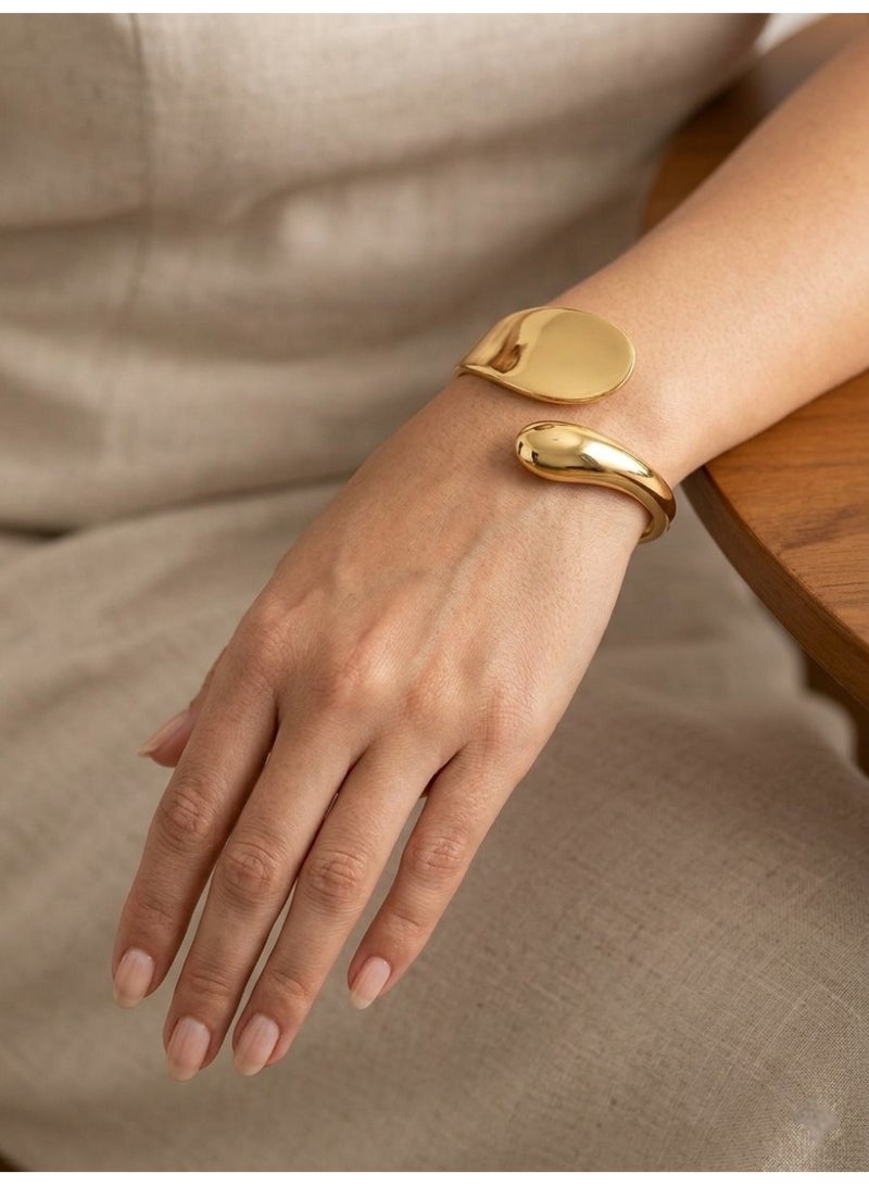A sophisticated open gold cuff bracelet featuring a unique sculpted organic design with asymmetrical ends—one flat and one smooth, rounded drop-like form—for a modern and distinctive look on your wrist